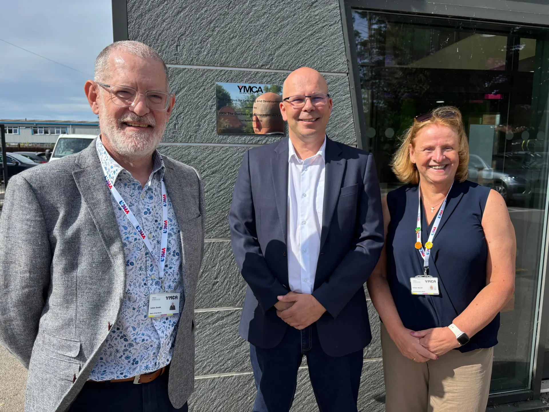 Julian Smith (Chair of the Board), incoming CEO Matt Jones and outgoing CEO Gillian Sewell standing outside the entrance to YMCA Derbyshire's Campus for Learning and Development on London Road, Derby, smiling for a photograph in bright sunshine.
